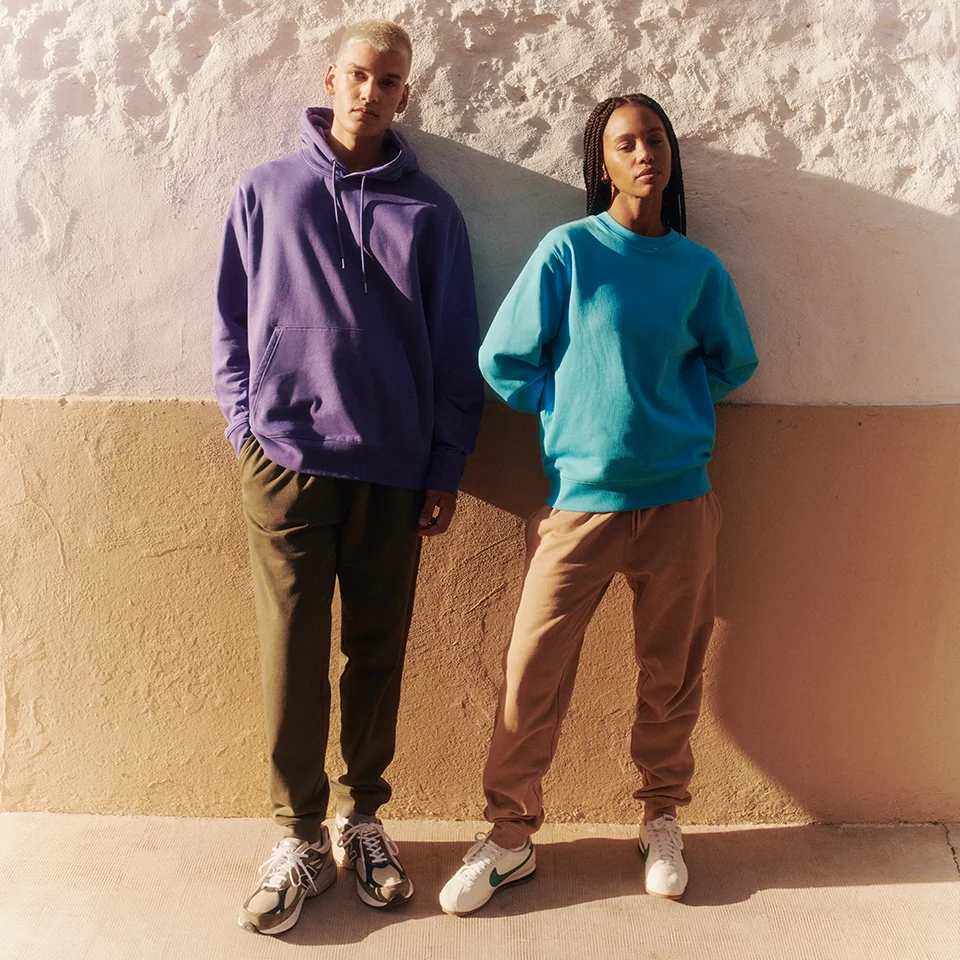 two fashionable models leaning against a wall wearing matching sweatpants and colourful hoodies